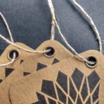 Tags of double-thick brown Kraft cover stock featuring a geometric design, reinforced with our antique brass eyelets and strung with our 3-ply hemp string.